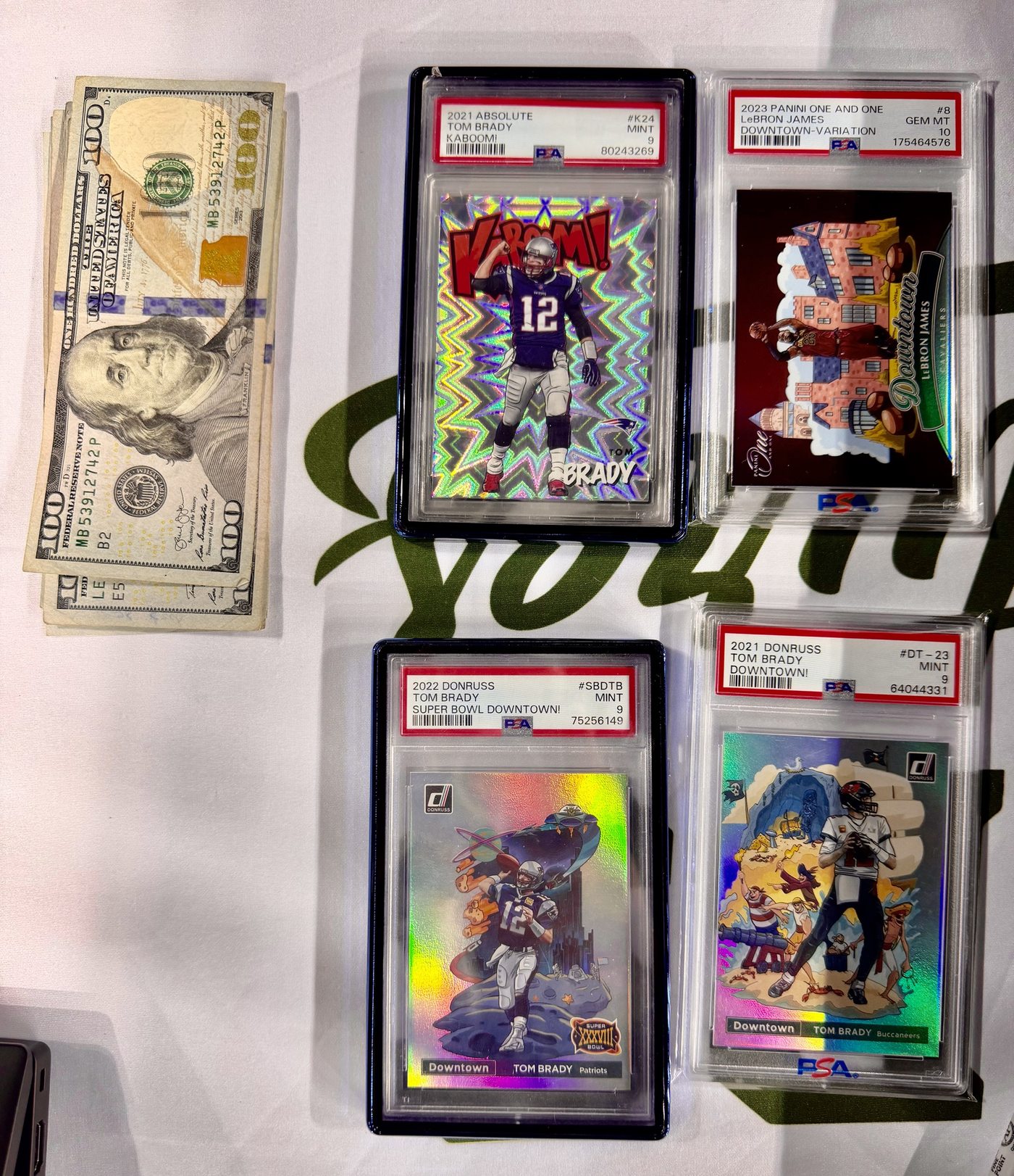 Four graded cards and cash on the Slabs & Signatures table at the Space City Card Show: a Tom Brady Kaboom PSA 9, a LeBron James Downtown variation PSA 10, and two Brady Downtowns